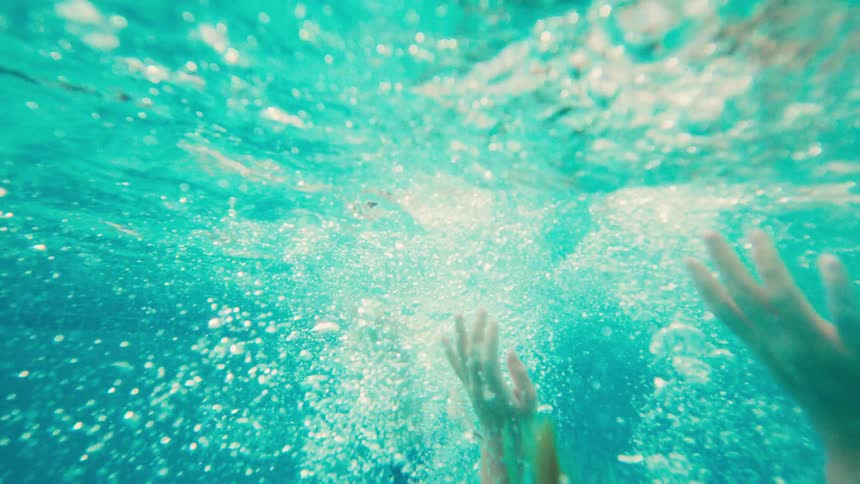 A slow-motion, underwater shot captures a woman in a swimsuit sinking into the turquoise water, her form becoming lost in the bubbles
