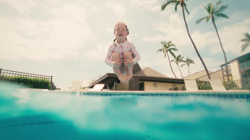 Slow motion underwater view of a joyful young girl doing a cannonball jump into a tropical resort swimming pool
