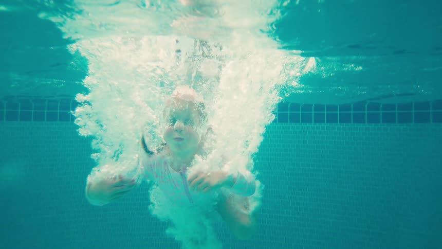 Slow motion underwater view of a joyful young girl doing a cannonball jump into a tropical resort swimming pool