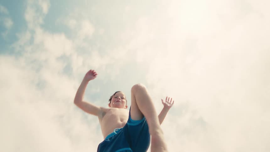 Dynamic low-angle slow-motion shot follows a young boy as he jumps into a swimming pool on a sunny day