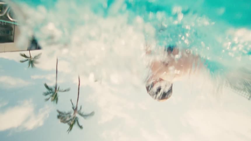 Dynamic low-angle slow-motion shot follows a young boy as he jumps into a swimming pool on a sunny day