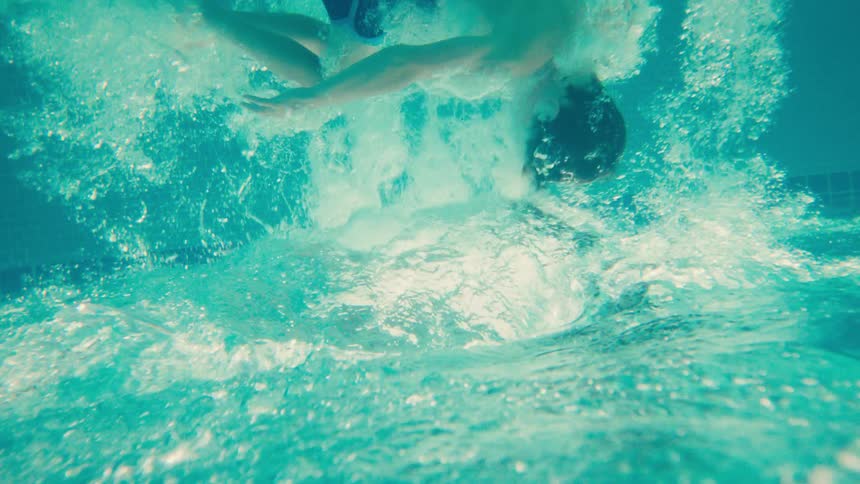 An underwater slow-motion view captures a young boy jumping into a swimming pool, creating a big splash and a flurry of bubbles