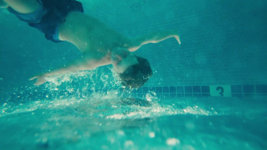 Slow motion underwater footage captures a boy floating serenely upside down in a turquoise swimming pool