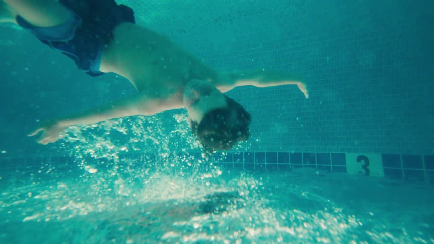 Slow motion underwater footage captures a boy floating serenely upside down in a turquoise swimming pool