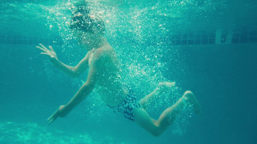 A young boy floats gracefully underwater in a sunlit swimming pool in a dreamy slow-motion shot