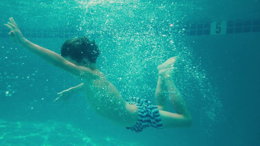 A young boy floats gracefully underwater in a sunlit swimming pool in a dreamy slow-motion shot