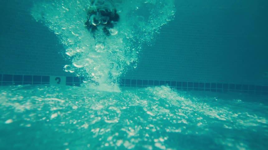 An upside-down, slow-motion, split-view shot captures a person with goggles splashing into a tropical resort swimming pool