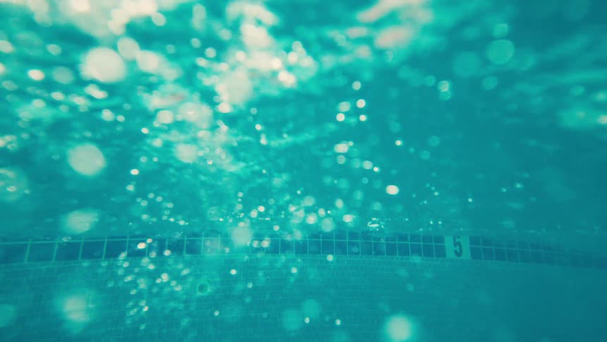 A dreamy, slow-motion underwater view of a turquoise swimming pool with sparkling light and bubbles rising to the surface