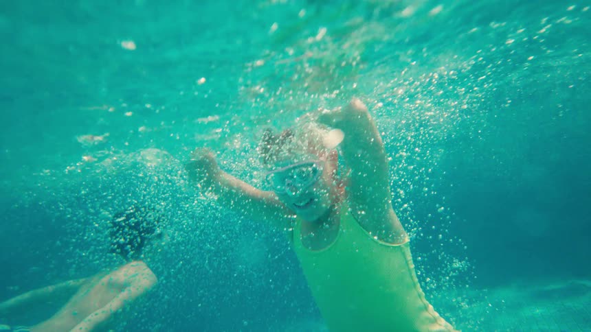 A slow-motion underwater shot shows a happy young girl in goggles smiling and waving at the camera while swimming in a pool with her brother