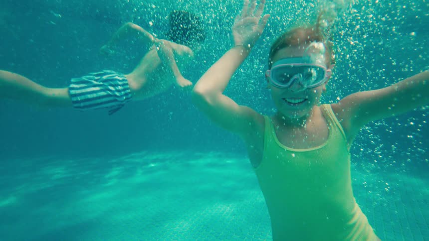 A slow-motion underwater shot shows a happy young girl in goggles smiling and waving at the camera while swimming in a pool with her brother