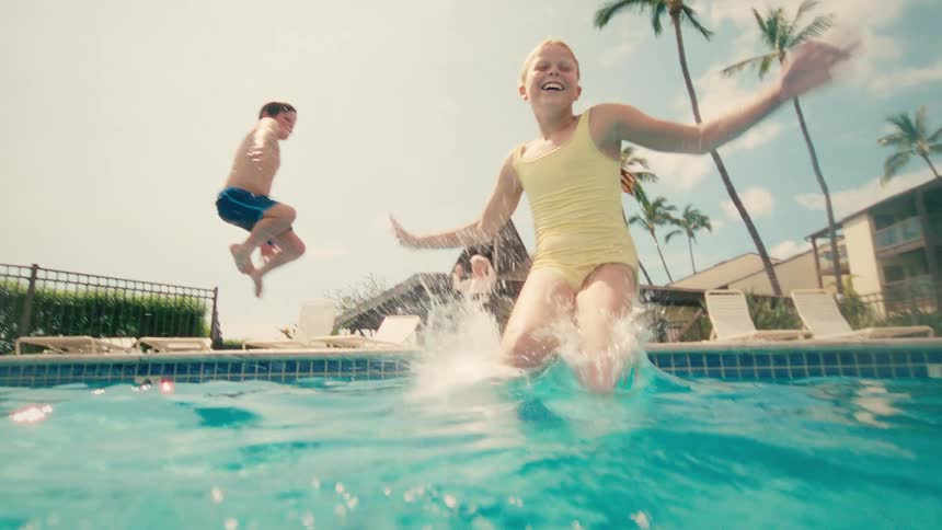 Kids joyfully jump into a swimming pool in slow motion, with the camera capturing the energetic splash from an underwater perspective