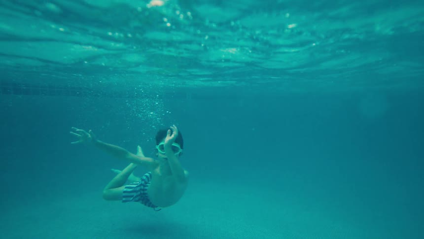 A young boy with swim goggles playfully waves at the camera while floating gracefully underwater in slow motion