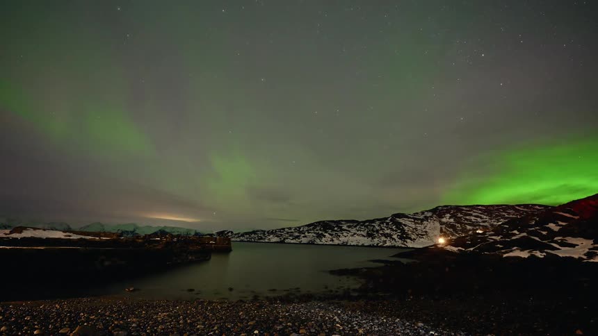 A stunning timelapse shows the vibrant green Northern Lights dancing across the starry night sky over a snowy, mountainous coastline