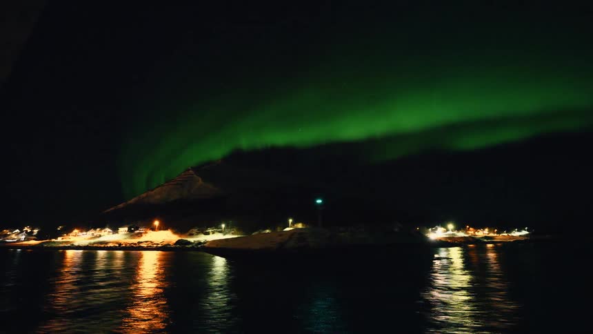 The magical green glow of the Northern Lights illuminates a snowy mountain and coastal village at night, with reflections on the water
