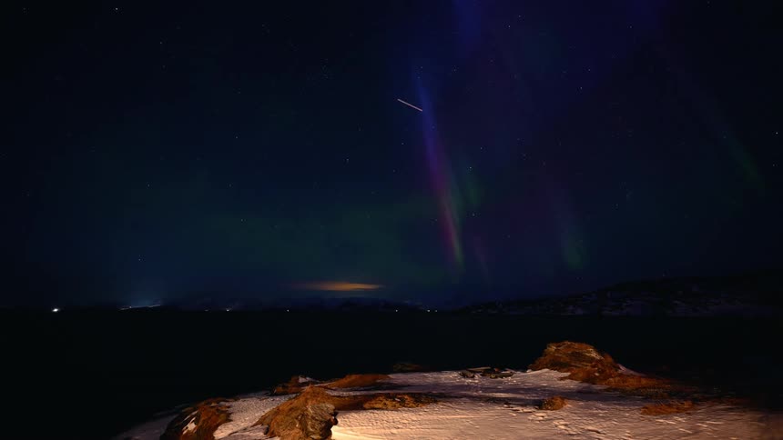 A stunning timelapse captures the vibrant green, blue, and purple Northern Lights dancing across the starry night sky over a snowy arctic landscape
