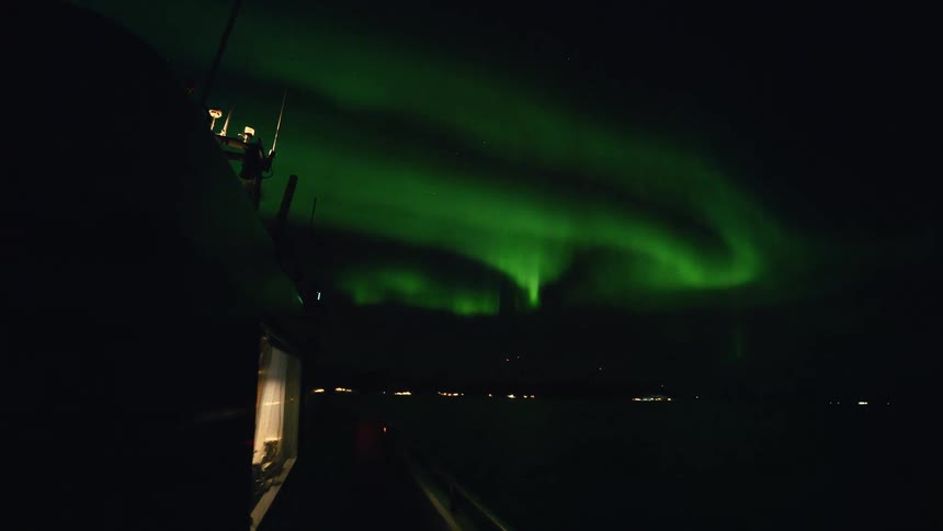 The vibrant green aurora borealis swirls and dances in the dark night sky, viewed from a boat cruising along a distant, illuminated coastline