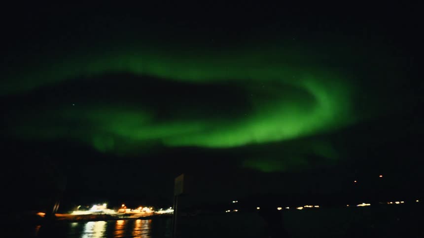 The beautiful green aurora borealis swirls and dances in the night sky above a snowy coastal town and harbor