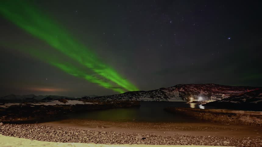 A vibrant green aurora borealis dances in a timelapse across the starry night sky over a snowy arctic bay and mountains
