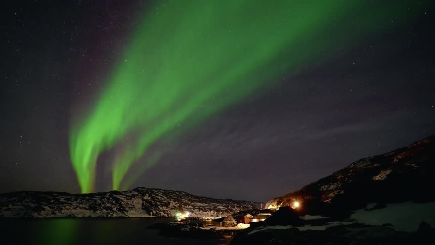 A stunning timelapse shows the vibrant green Aurora Borealis dancing across the starry night sky over a snowy mountain village by the water