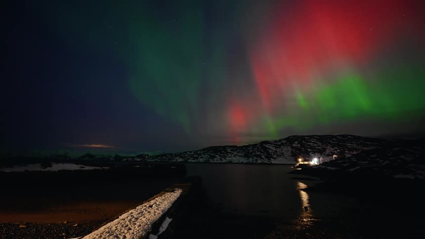 A breathtaking timelapse captures the vibrant green and red dance of the Aurora Borealis over a tranquil, snowy fjord at night