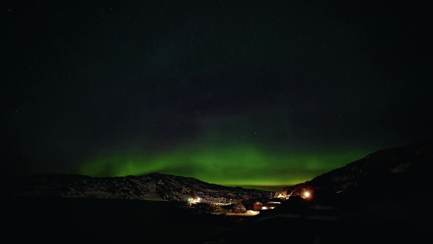 A stunning timelapse shows the vibrant green Aurora Borealis dancing in the starry night sky over a small coastal village nestled in snowy mountains