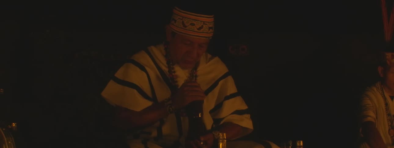 An indigenous spiritual leader in traditional clothing performs a solemn ritual in a dark, candlelit setting