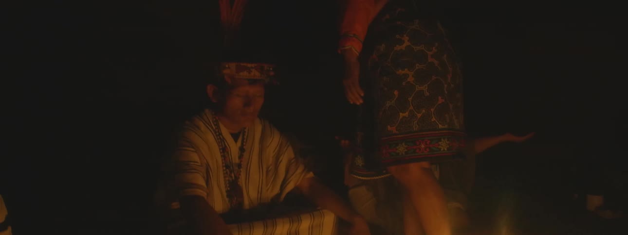 A group of indigenous people dressed in traditional clothing participate in a solemn candlelit ceremony in the dark