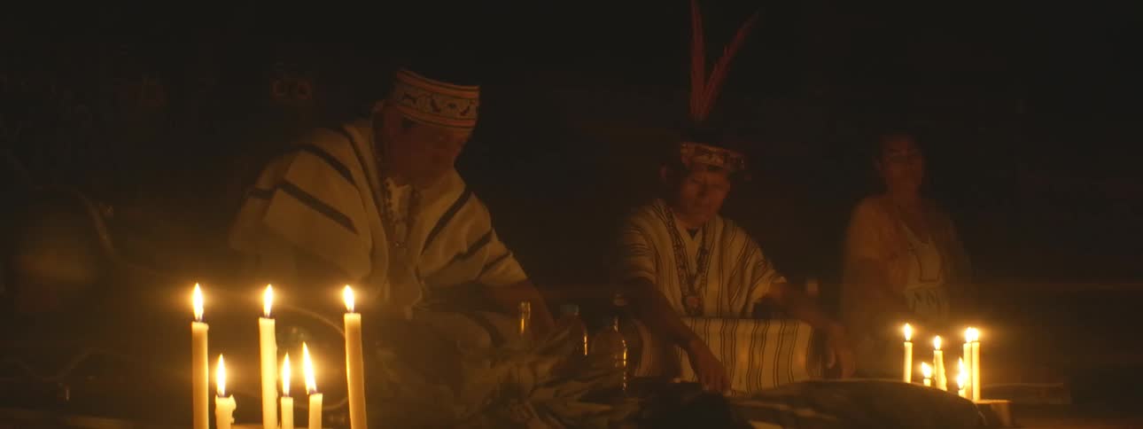 Indigenous shamans wearing traditional garments and headdresses conduct a sacred candlelit ceremony in a dark room