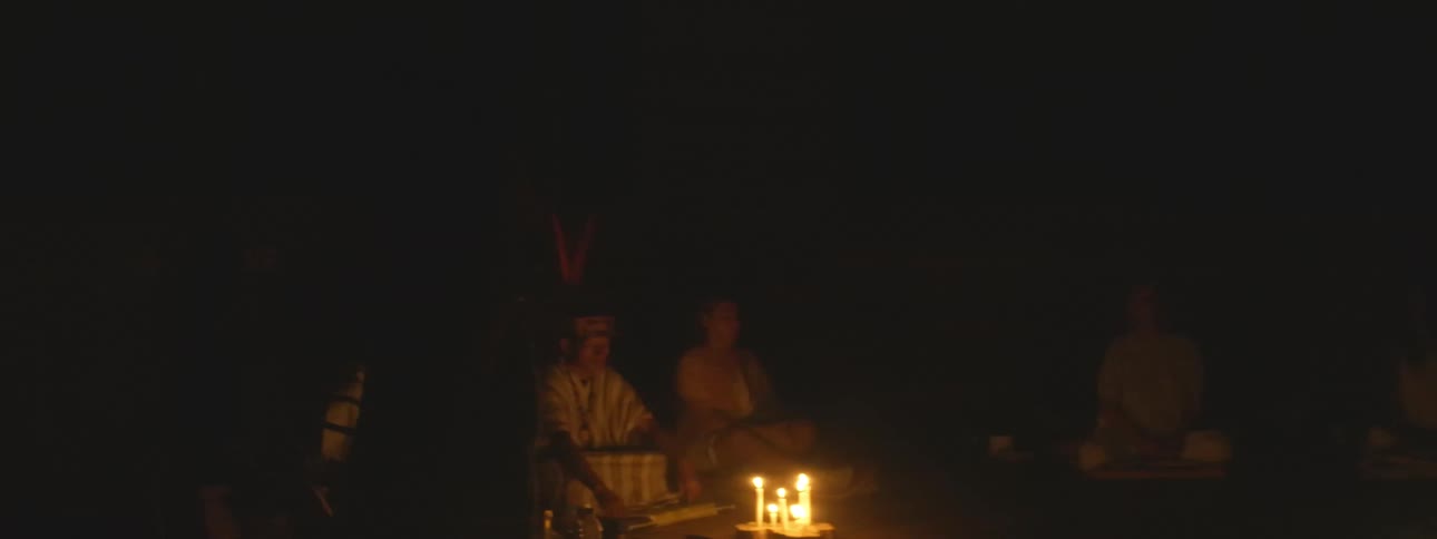 A group of people participate in a sacred shamanic ceremony in a dark room illuminated only by flickering candlelight