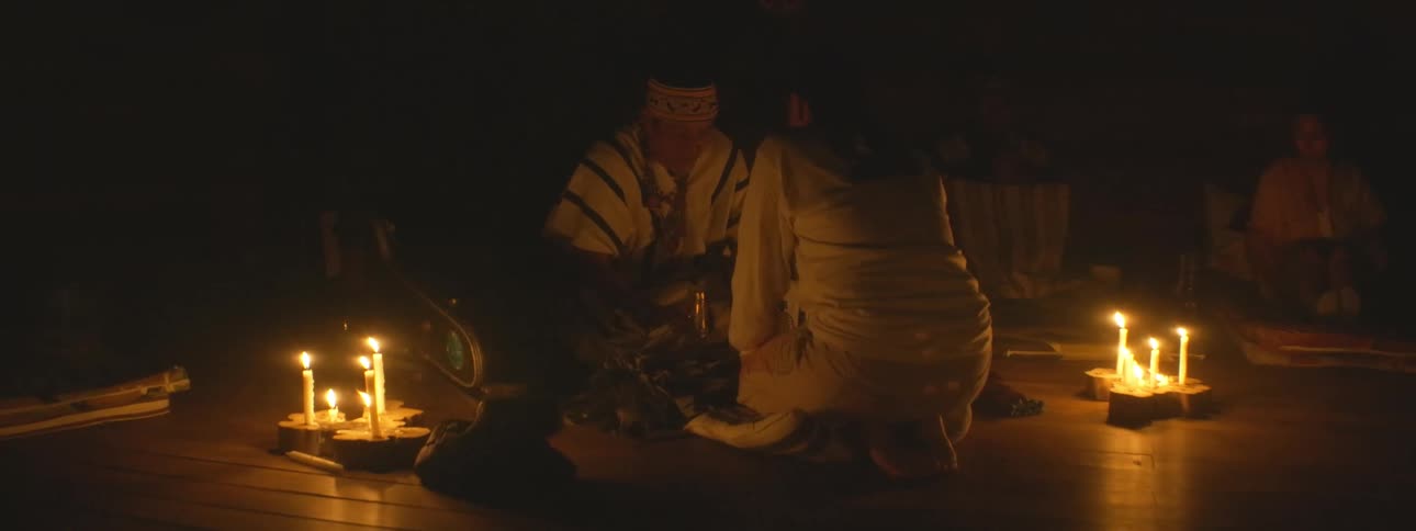 A shaman in traditional attire performs a sacred healing ceremony for a woman in a dark room illuminated by flickering candlelight