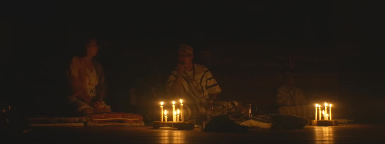 A shaman in traditional clothing leads a spiritual ceremony with other participants in a dark room illuminated only by flickering candles