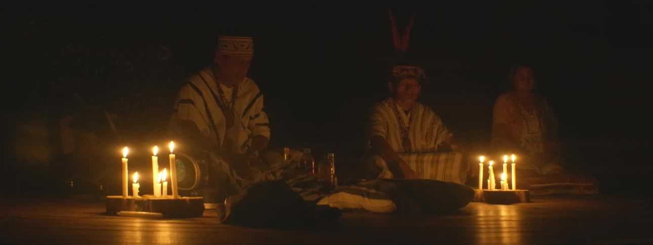 Three indigenous shamans in traditional attire sit on the floor in a dark room, illuminated by candlelight during a sacred spiritual ceremony