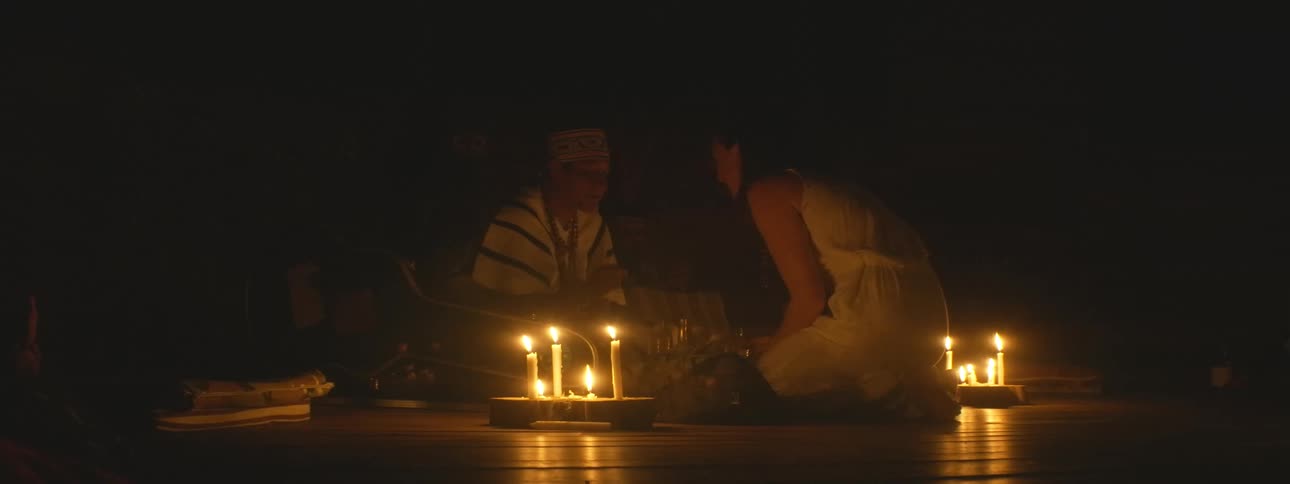A shaman and a woman participate in a traditional spiritual ceremony illuminated by flickering candles in a dark room
