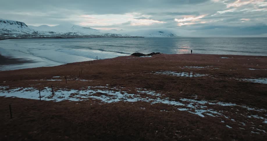 A majestic aerial shot slowly approaches a lone person standing on a rugged, snowy coastline overlooking the ocean and distant mountains