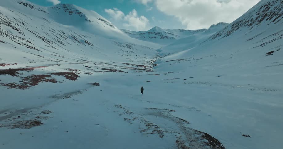 An aerial shot follows a lone hiker walking through a vast, snow-covered valley surrounded by mountains