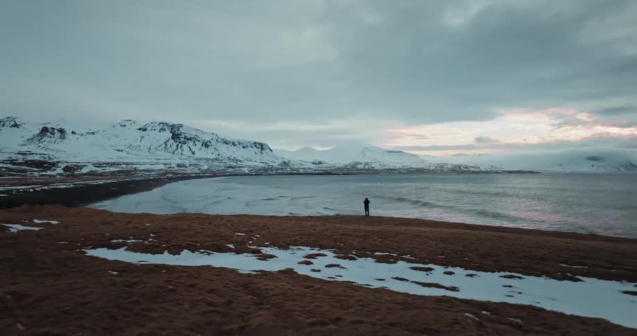 An aerial shot pulls back to reveal a solitary figure standing on a grassy, snow-dusted cliff, overlooking a vast bay and snow-covered mountains at dusk