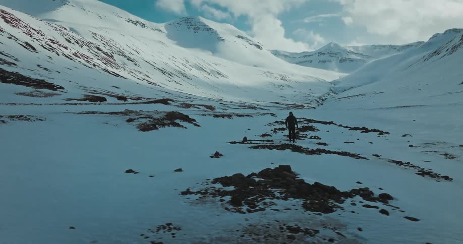 An aerial view follows a lone hiker as they walk through a vast and beautiful snow-covered mountain valley