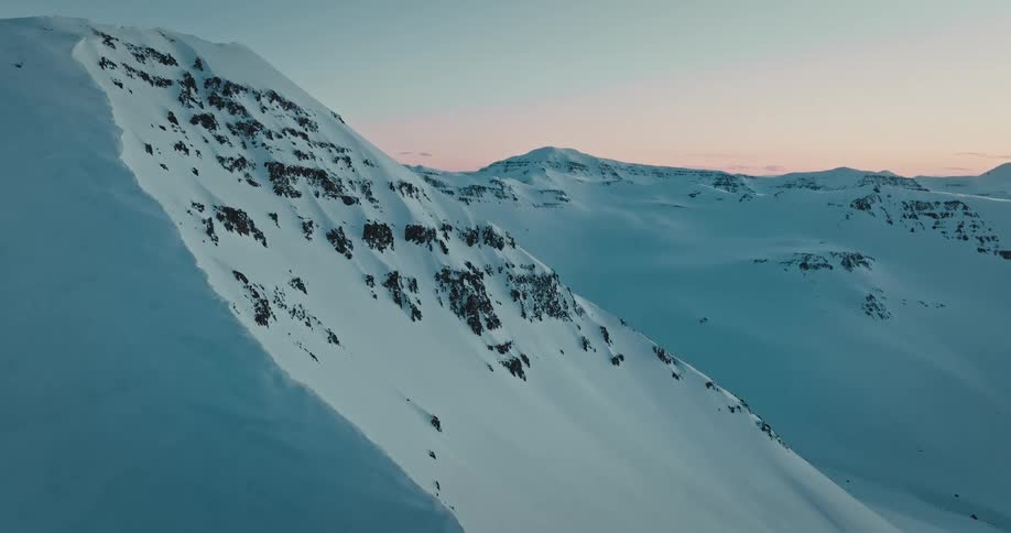 Aerial drone footage reveals a vast, serene snow-covered mountain range during a tranquil winter sunset