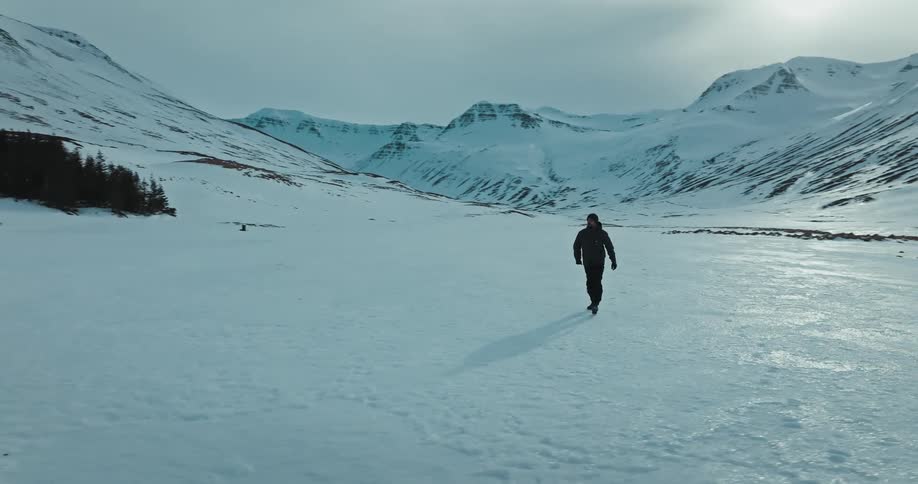 Cinematic aerial shot follows a man walking alone through a vast, frozen mountain valley in Iceland