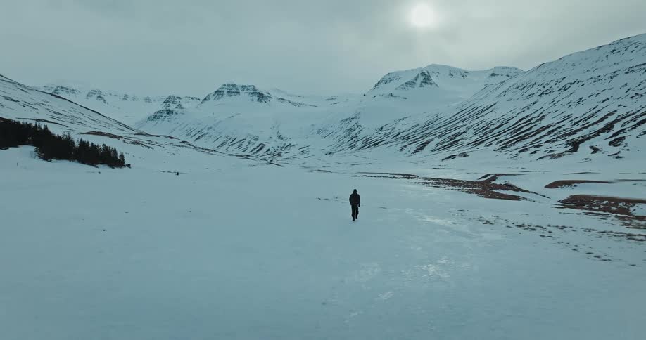 An aerial view follows a solitary figure walking through a vast, snow-covered mountain valley on a cold, overcast day