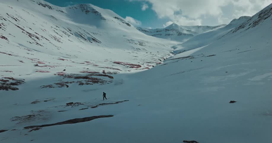 A lone hiker makes their way across a vast, sun-dappled, snow-covered mountain valley in a stunning aerial shot