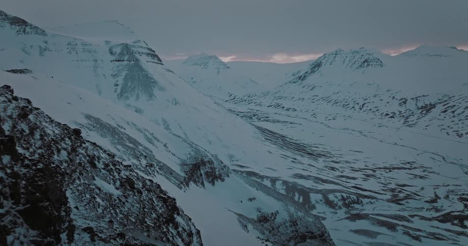 A dramatic aerial shot flies past a dark cliff face to reveal a stunning, snow-covered mountain valley landscape at dusk