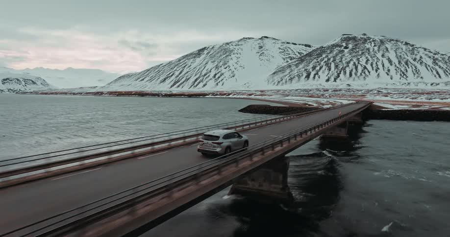 A silver SUV drives across a bridge over the water, surrounded by a dramatic, snow-covered mountain landscape in a moody aerial shot