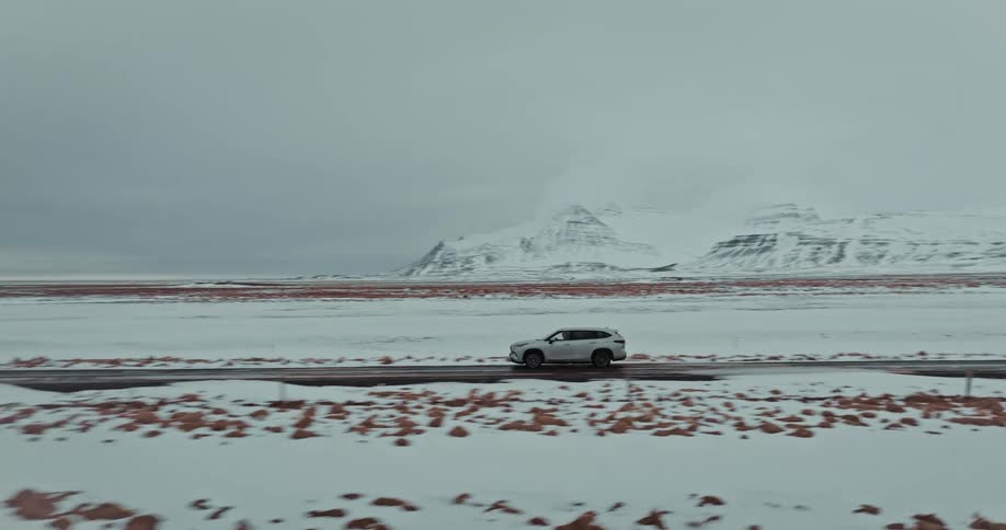 Aerial footage tracks a silver SUV driving along a remote road through a vast, snow-covered landscape with majestic mountains on the horizon