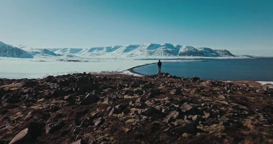 A solitary figure stands on a rocky promontory overlooking a vast, snow-covered landscape and a serene blue fjord, as an aerial camera pulls back to reveal the epic scale