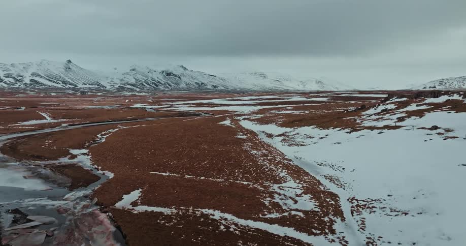 Aerial view rises over a vast, frozen Icelandic tundra with a winding river and snow-capped mountains in the distance