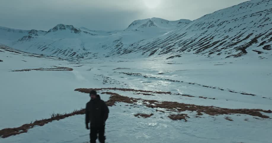 Aerial view follows a lone hiker walking through a vast snowy mountain valley, revealing the expansive frozen landscape