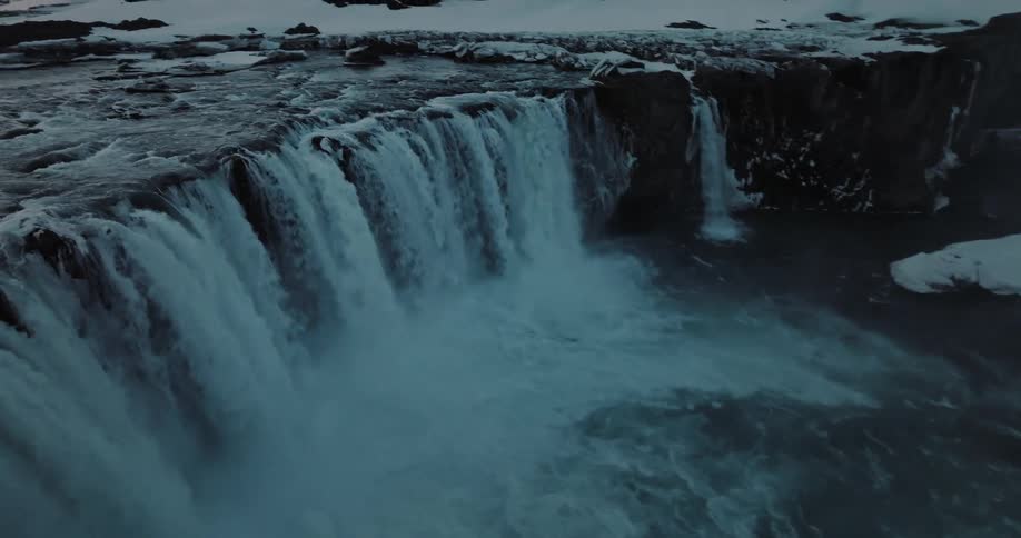 A stunning high-angle view reveals the immense power of a wide waterfall crashing into a river amidst a bleak, snowy winter landscape