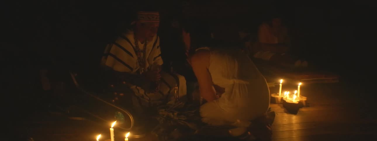 An indigenous shaman performs a traditional spiritual blessing ceremony on a kneeling woman in a dark, candlelit room