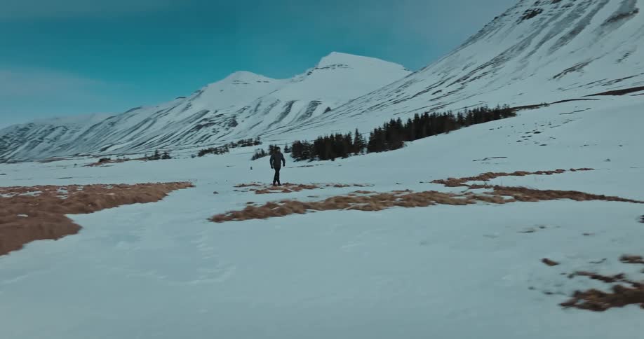 A tracking shot follows a lone hiker walking through a vast, snow-covered mountain valley